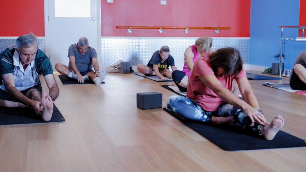 A group of seniors and adults participating in a yoga or stretching class on mats in a gym studio, performing seated head-to-knee forward folds.