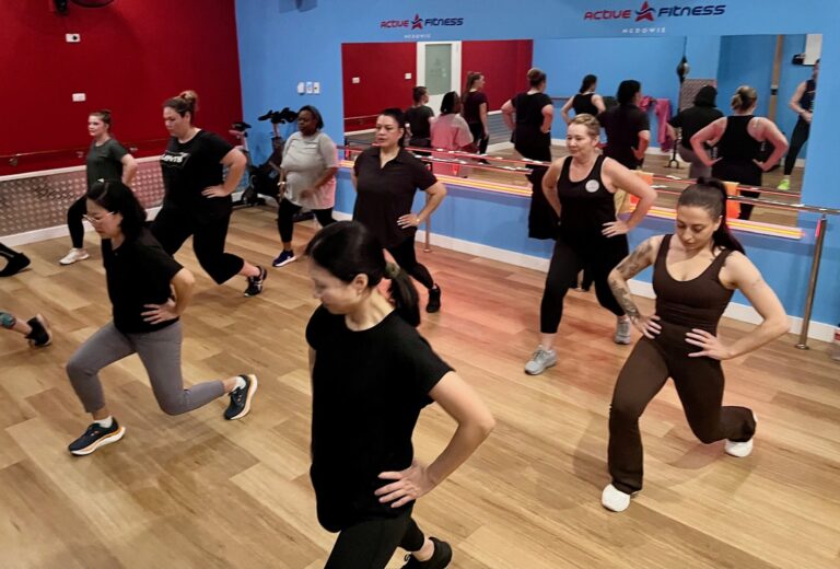 A group of women in athletic wear perform reverse lunges with their hands on their hips during a fitness class.