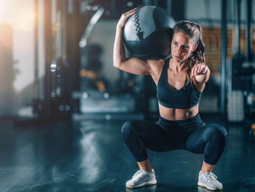 A muscular woman performs a deep squat while holding a heavy medicine ball on one shoulder in a gym setting.