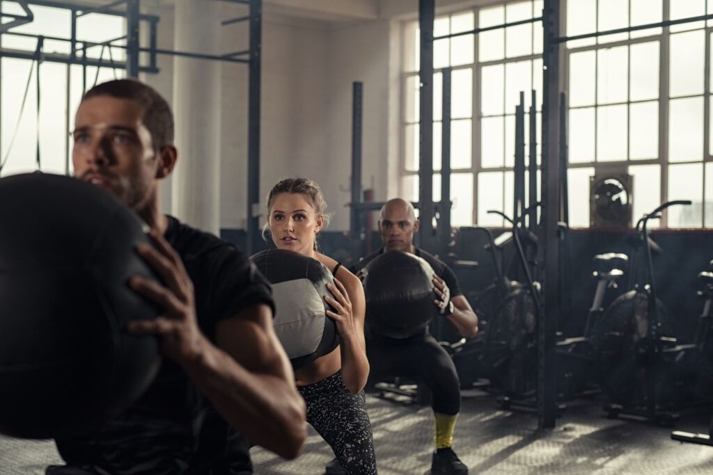 A group of athletes in a CrossFit-style gym perform squats while holding heavy medicine balls in a "goblet" position.