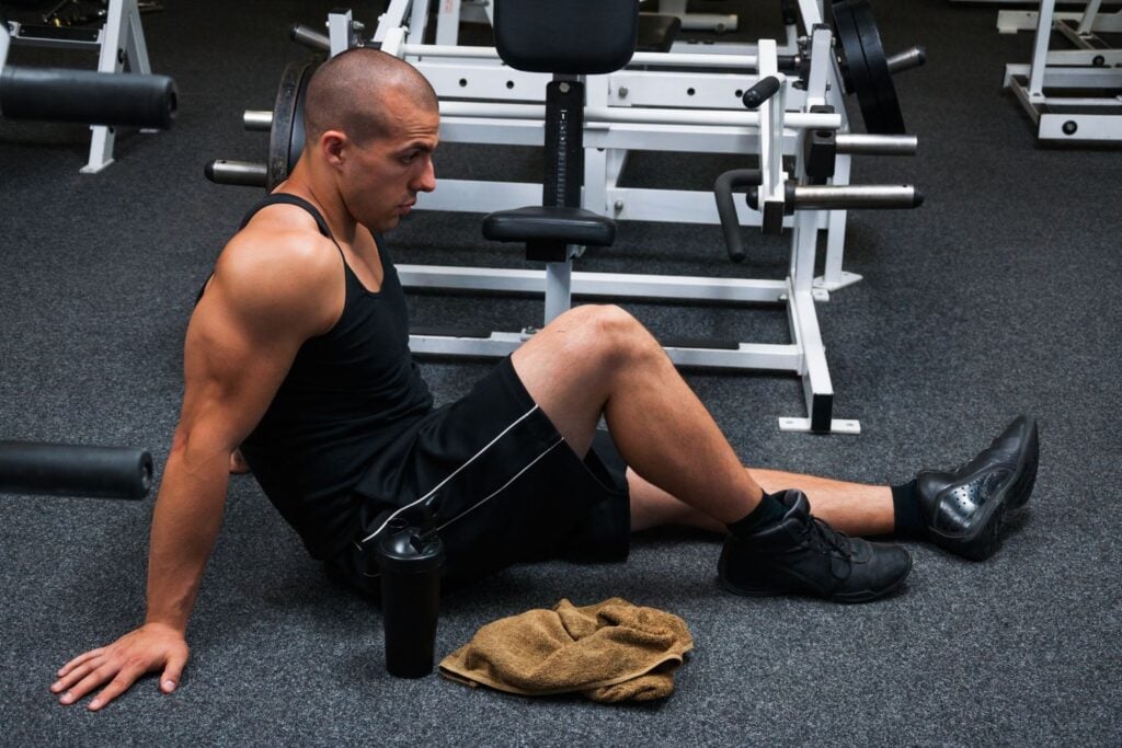 A muscular man in a black tank top sits on the gym floor taking a rest
