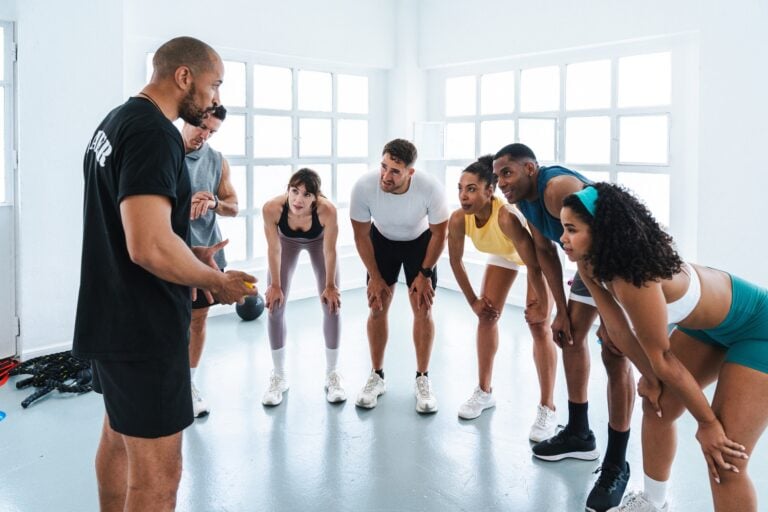 A fitness trainer addressing a small group of focused participants leaning forward during a workout session.