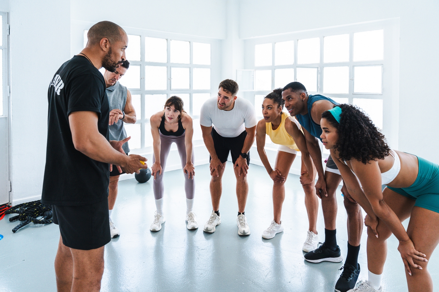 A fitness trainer addressing a small group of focused participants leaning forward during a workout session.