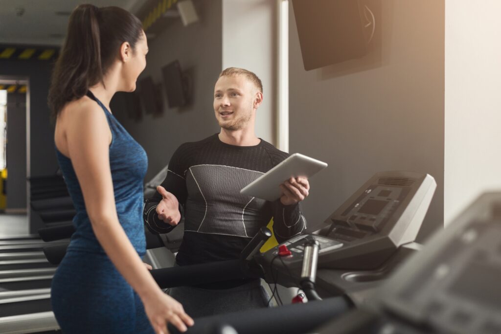 A male personal trainer holding a tablet and talking to a woman on a treadmill during a consultation in a modern gym