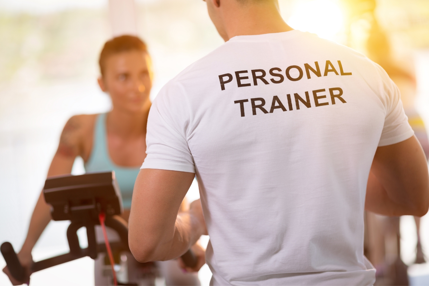 A personal trainer in a white t-shirt seen from behind, guiding a woman on an exercise bike in a sunlit gym.
