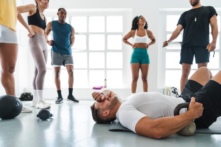 An exhausted man lies on a gym floor after a workout, surrounded by supportive gym members and a trainer