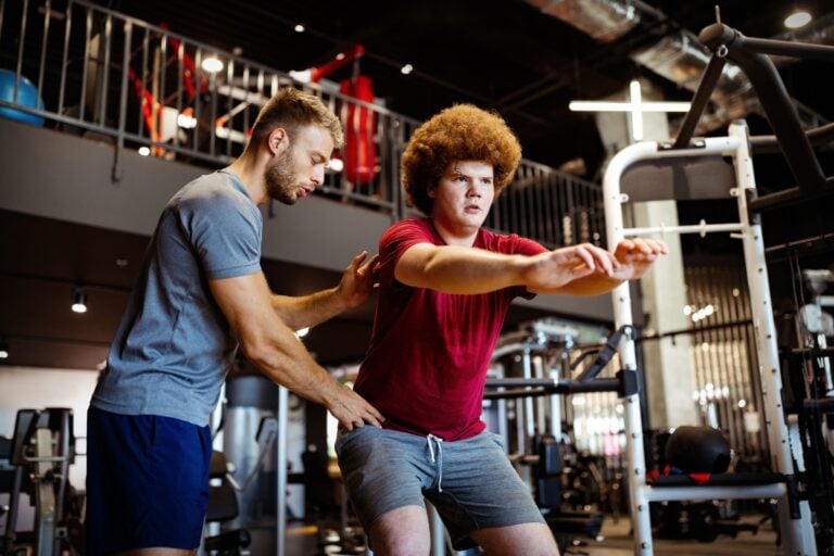A trainer and client working together inside a well-equipped gym, trainer observing or correcting form, natural and candid rather than posed. Clean, bright space with visible equipment in the background.
