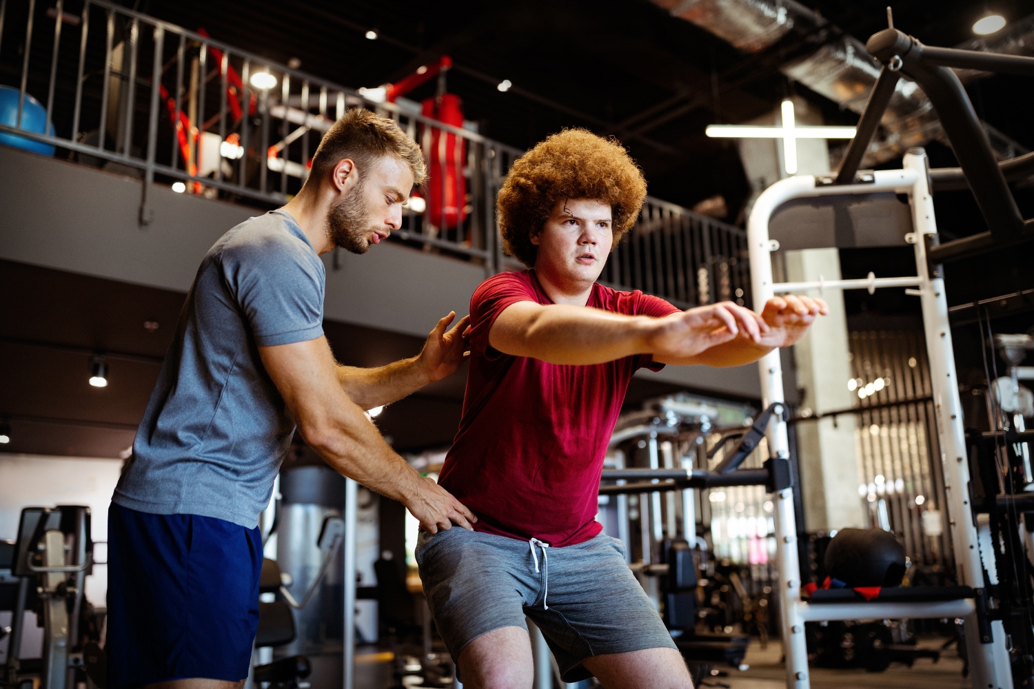 A trainer and client working together inside a well-equipped gym, trainer observing or correcting form, natural and candid rather than posed. Clean, bright space with visible equipment in the background.