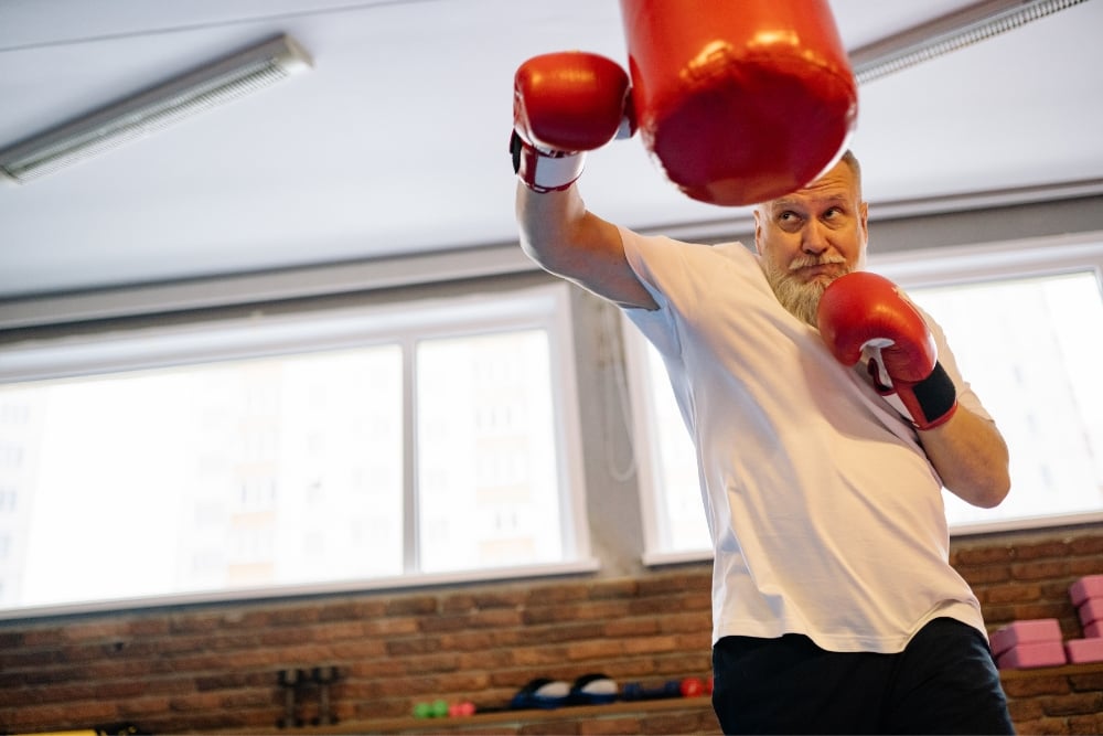A low-angle shot of an older man with a grey beard wearing red boxing gloves and a white t-shirt, throwing an upward punch at a red heavy bag in a gym.