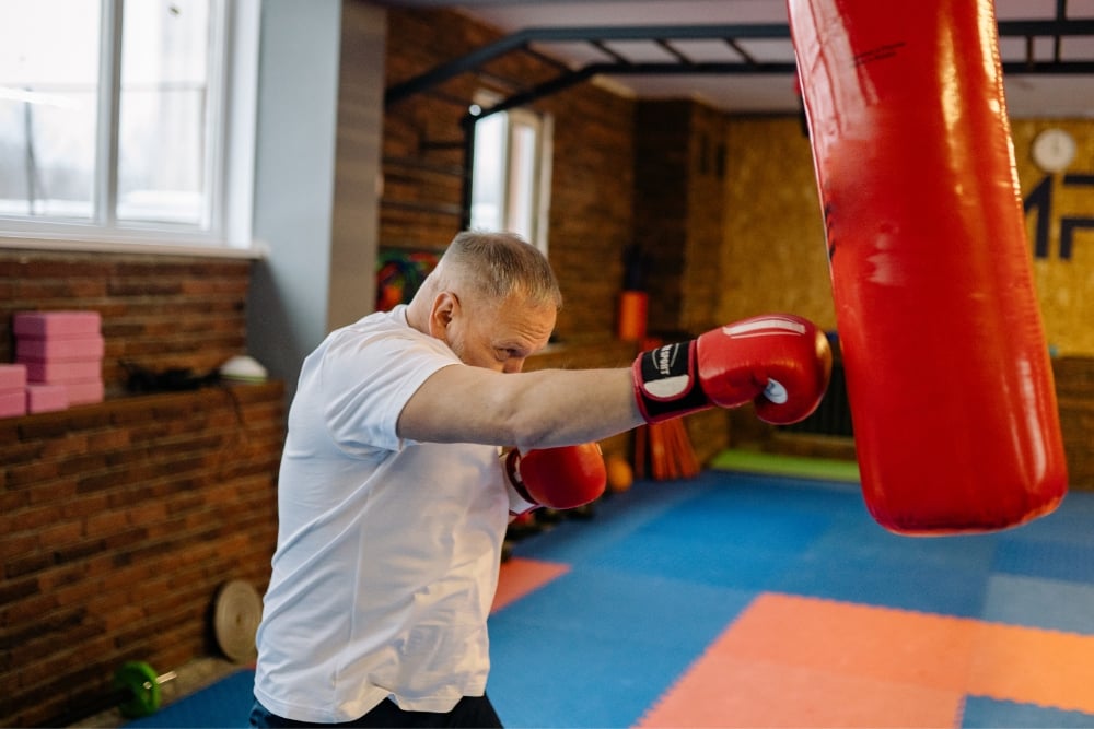 An older man in a white t-shirt and red boxing gloves throws a punch at a large red heavy bag in a gym with blue and orange floor mats.