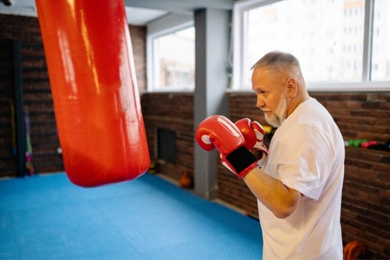 An older man with a white beard wearing boxing gloves and a white t-shirt stands in a gym, facing a large red punching bag.
