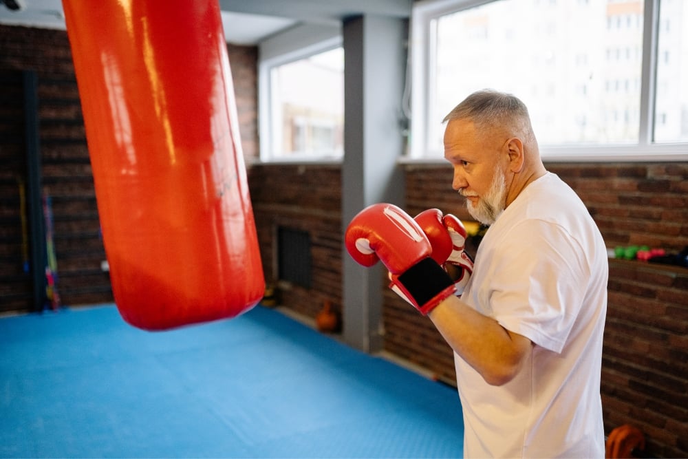 An older man with a white beard wearing boxing gloves and a white t-shirt stands in a gym, facing a large red punching bag.
