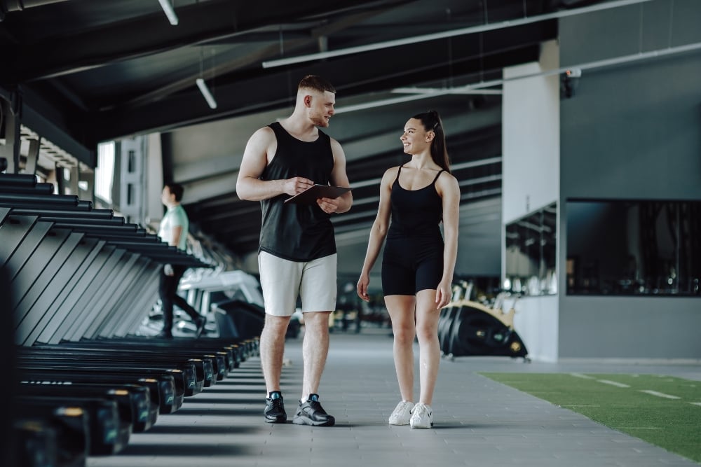 A male personal trainer holding a clipboard stands talking and smiling with a female client in black athletic wear.