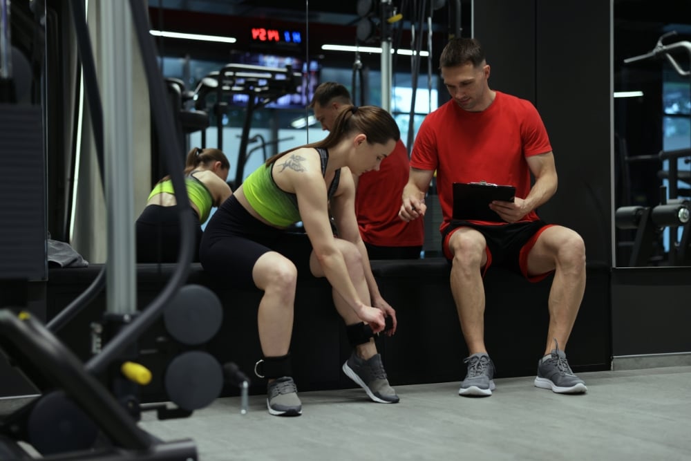 A female client sitting on a gym bench and adjusting an ankle strap weight while a male personal trainer sits beside her holding a clipboard.