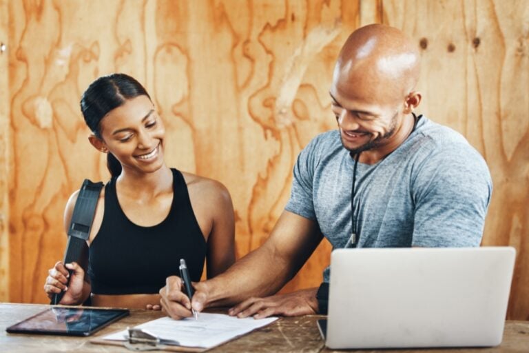 A smiling fitness trainer signs a document on a clipboard for a happy female client at a gym reception desk with a laptop and tablet.