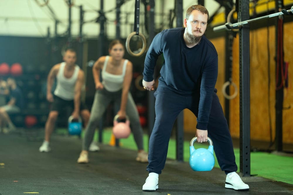 A man with a beard wearing dark sweats stands in a gym lifting a blue kettlebell with one hand during a group fitness class.