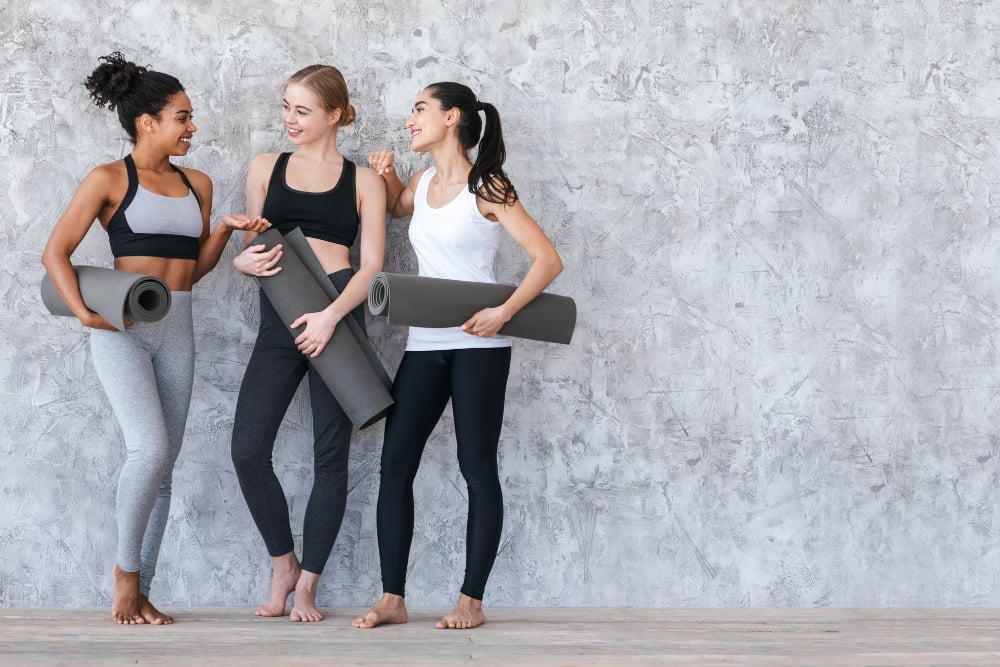 A three women in athletic wear standing together against a textured grey wall, holding rolled-up grey fitness mats and smiling while talking.
