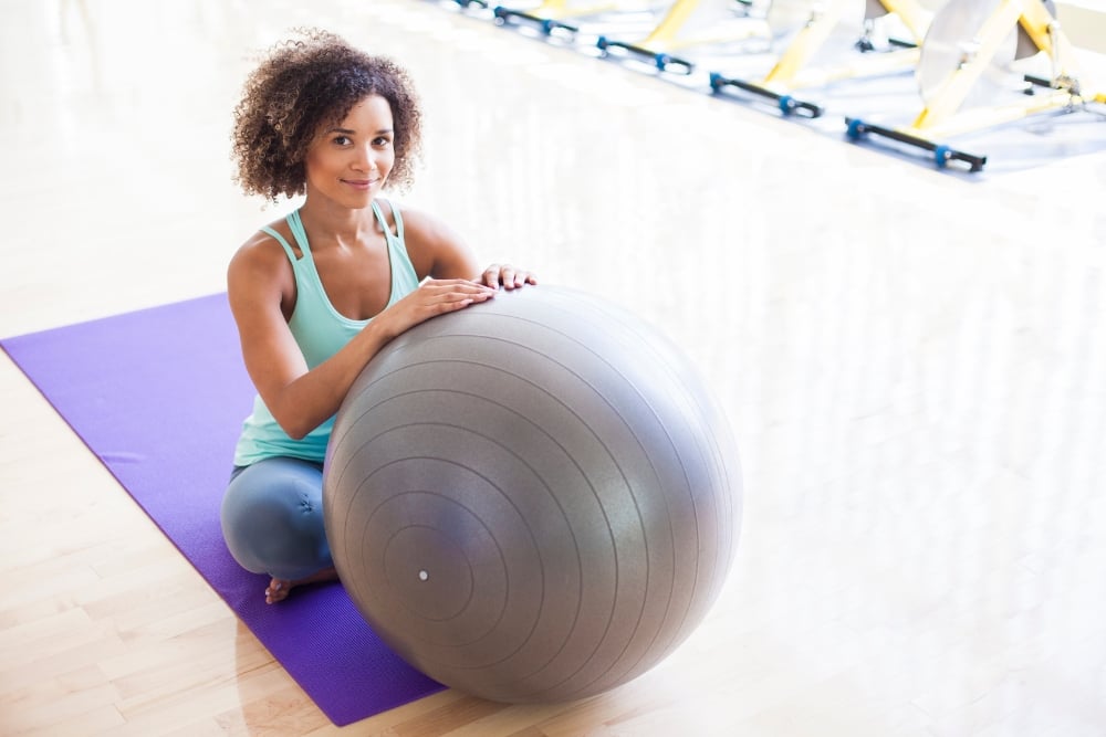 A woman with curly hair sitting cross-legged on a purple yoga mat in a gym, smiling and leaning against a large grey exercise ball.