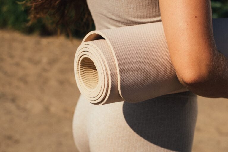 A close-up view of a person holding a rolled-up beige fitness mat under their arm.