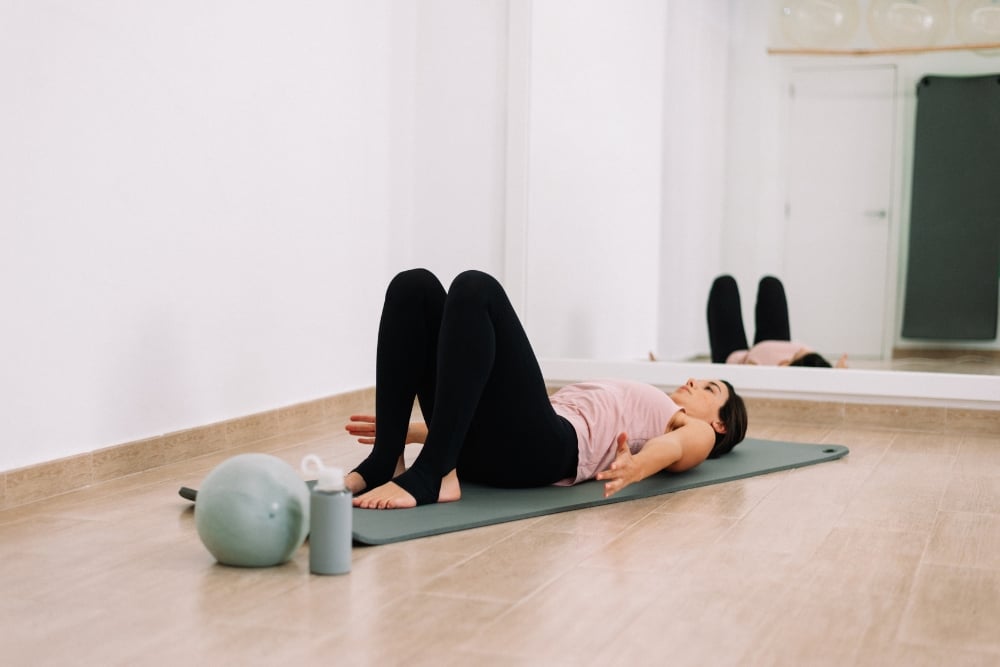 A woman lying on a yoga mat in a studio with her knees bent, next to a small exercise ball and water bottle.