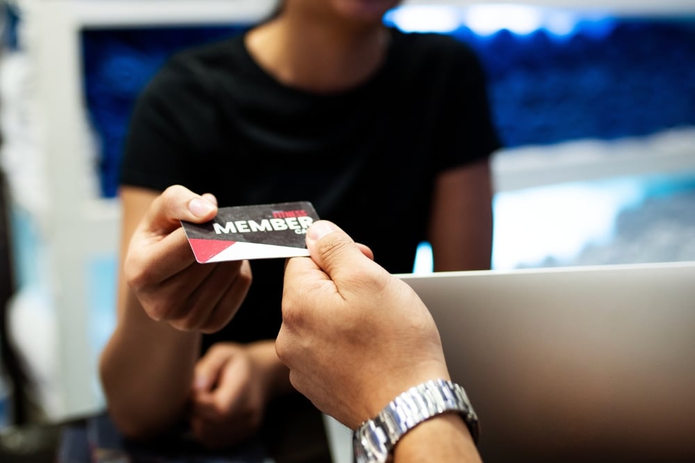 A close-up of a person handing over a "Fitness Member" card to a gym staff member at a reception desk.
