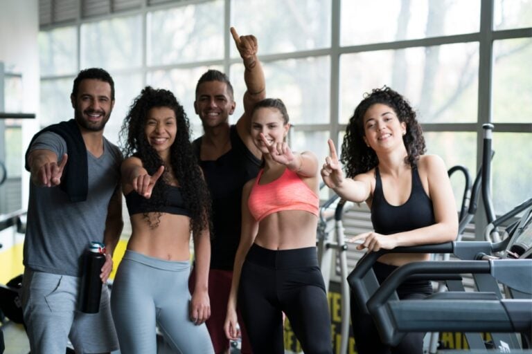 A group of five smiling friends in athletic wear posing together and pointing forward in a bright gym setting.