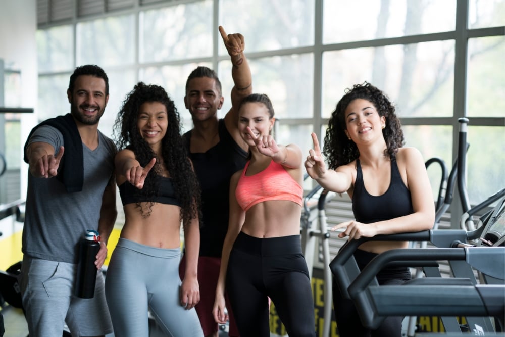 A group of five smiling friends in athletic wear posing together and pointing forward in a bright gym setting.