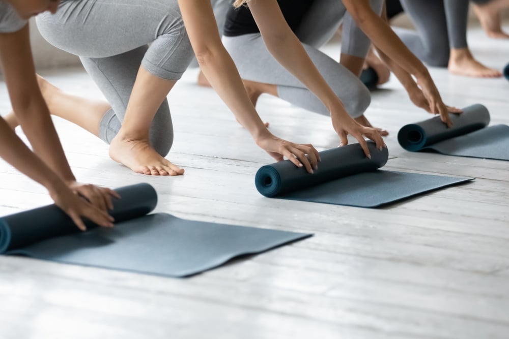 A row of people kneeling on a light wooden floor, each unrolling or rolling up a dark blue fitness mat.