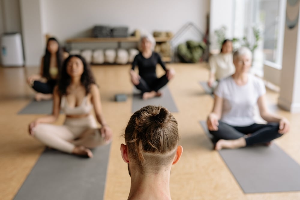 Group of people sitting cross-legged on mats in a quiet mat Pilates class led by an instructor.