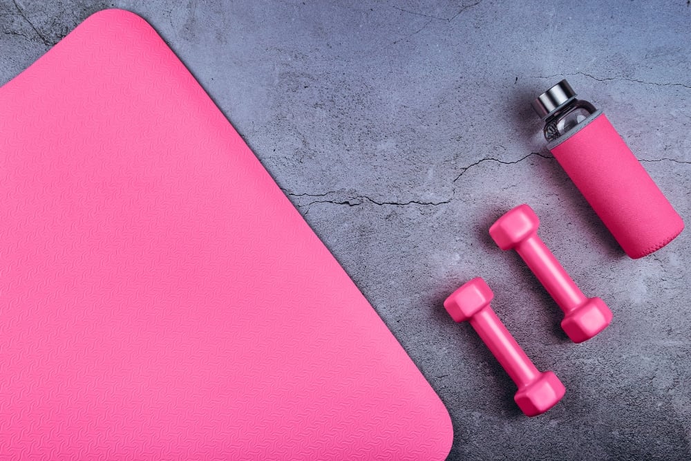 A flat lay view of fitness equipment, featuring a pink exercise mat, two pink dumbbells, and a pink-covered water bottle on a textured grey background.