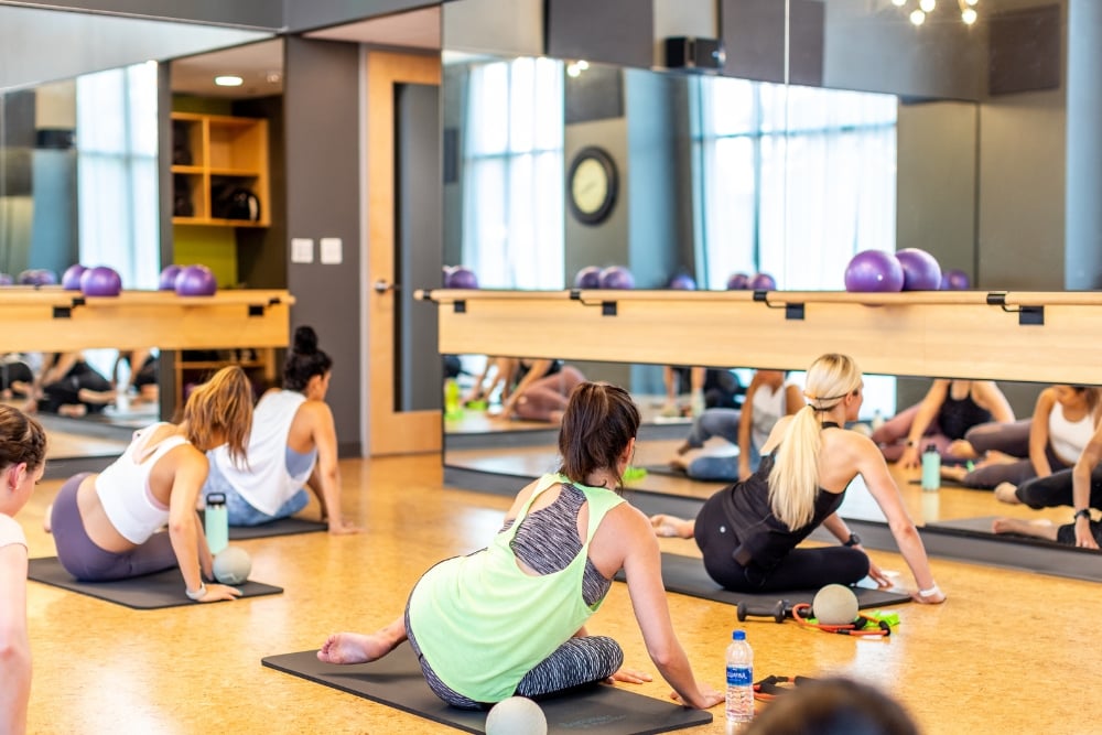 A group of women in a barre studio sitting on exercise mats while performing a seated core stretch facing mirrored walls.