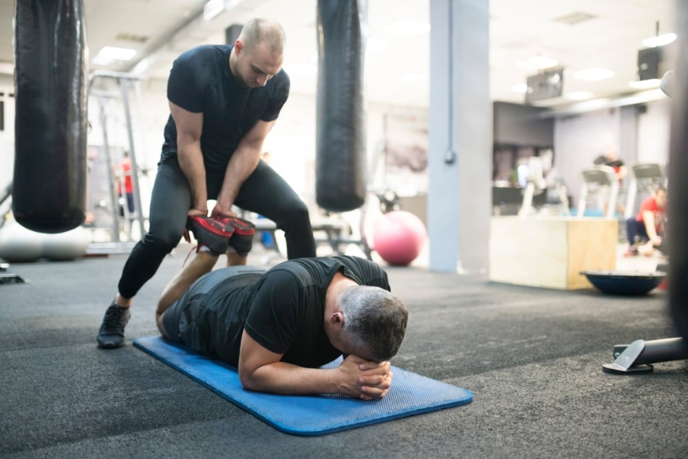 A trainer assisting a man lying on an exercise mat with a assisted leg stretch in a gym setting.