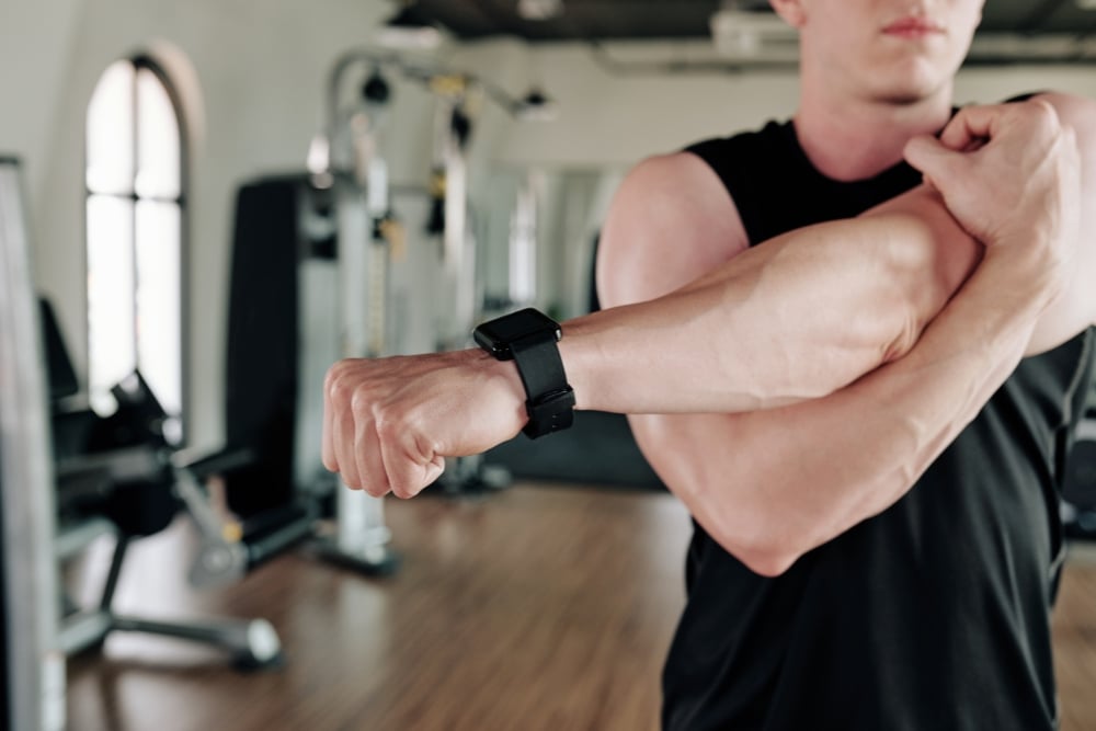 A close-up of a man wearing a smartwatch stretching his arm across his chest in a gym.