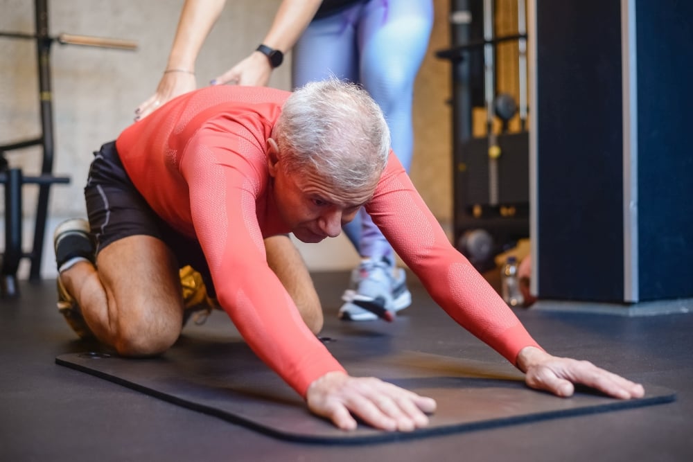 An older man in a red shirt performing a child's pose stretch on a mat with an instructor supporting his lower back.