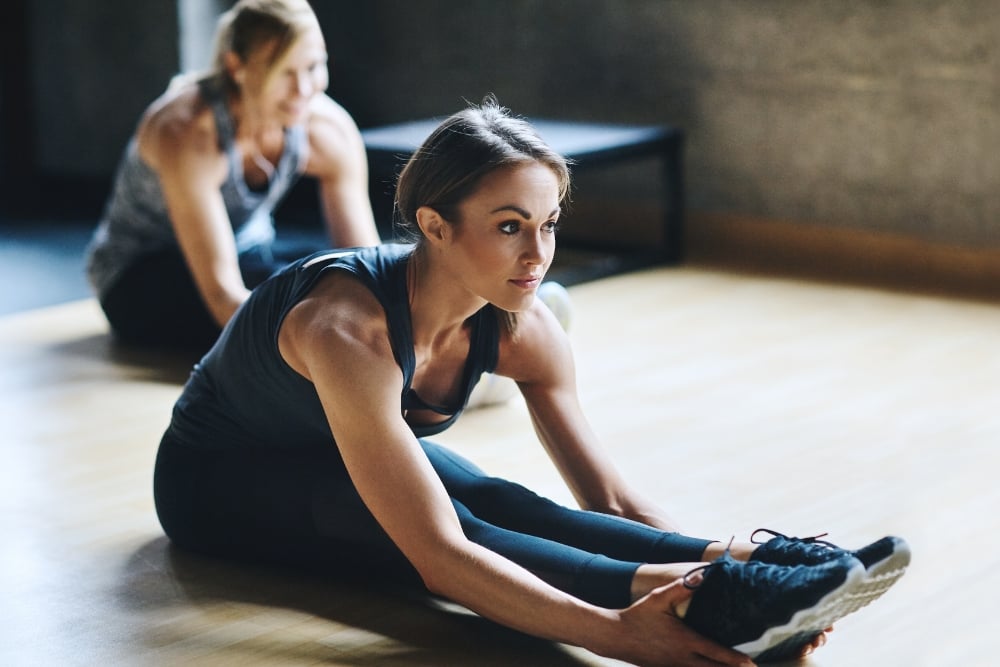 Woman sitting on gym floor doing a seated forward fold stretch and touching her feet.