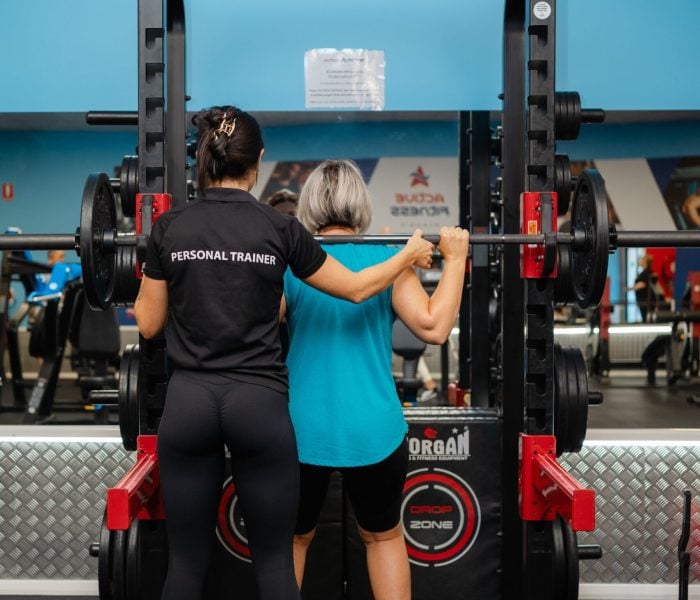 A personal trainer in a black shirt assists a woman in a blue tank top with a barbell squat inside a power rack at Active Fitness Medowie.