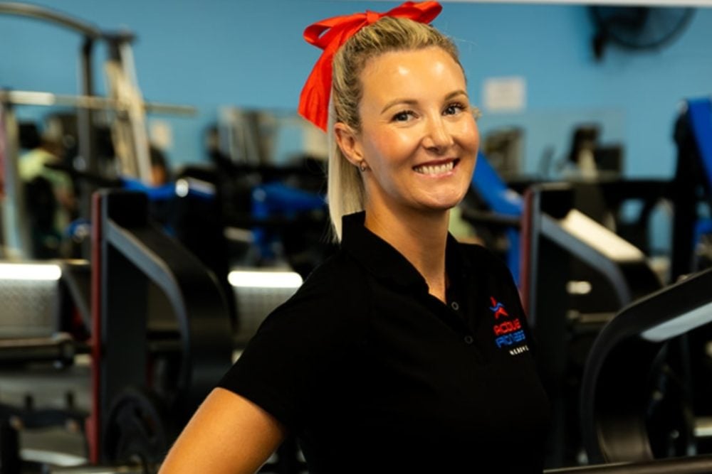 A smiling woman with a red bow in her hair, wearing a black polo shirt and blue shorts, stands with her hands on her hips in a gym.