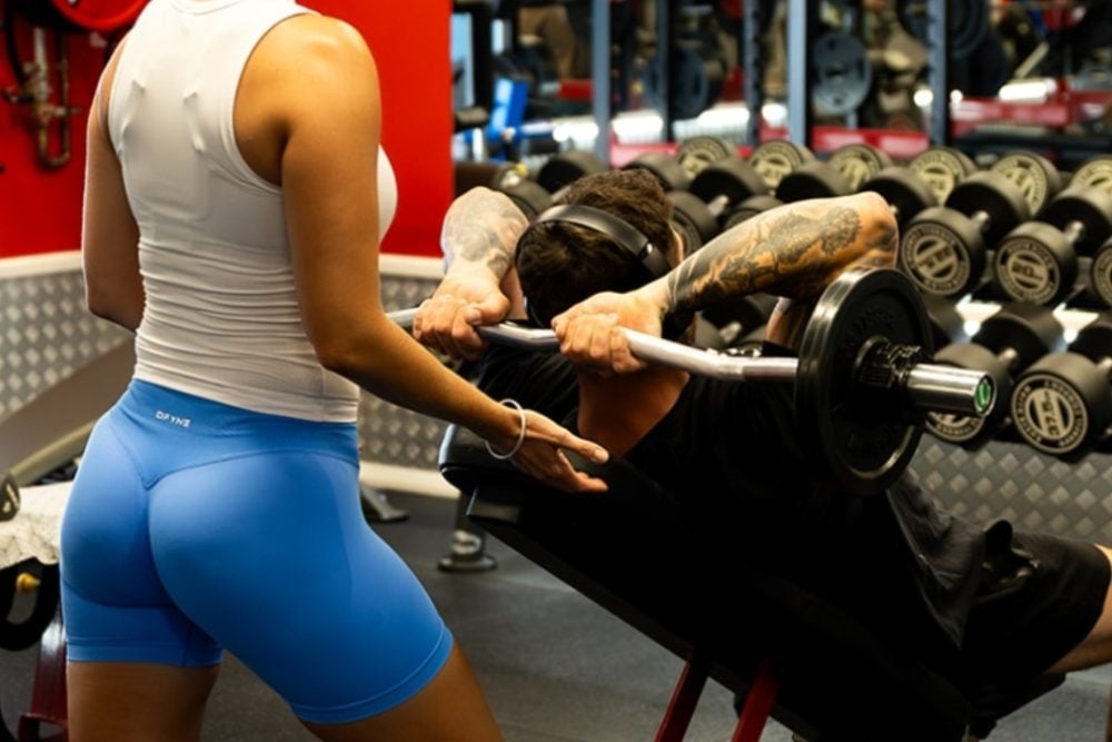 A woman in a white tank top and blue shorts spots a man performing skull crushers with an EZ bar on an incline bench at the gym.
