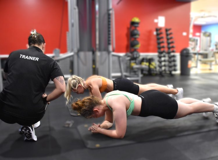 A fitness trainer in a black shirt watching two women performing planks on mats in a gym with red walls.