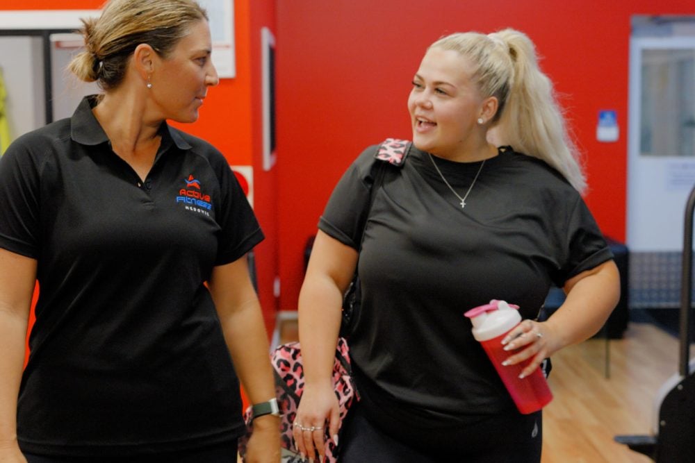 Two women talking at a gym, one wearing a staff polo shirt and the other holding a protein shaker bottle.