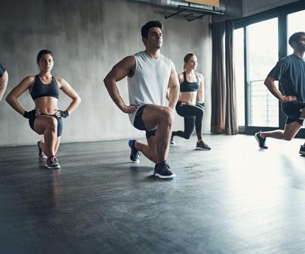 A group of people perform forward lunges together in a spacious, modern gym studio.