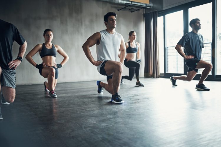 A group of people perform forward lunges together in a spacious, modern gym studio.