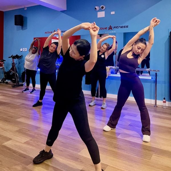 A group of women in athletic wear perform overhead side stretches together during a fitness class in a blue-walled studio.